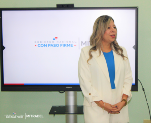 A woman in a white blazer stands in front of a screen displaying a government logo and the phrase "CON PASO FIRME."