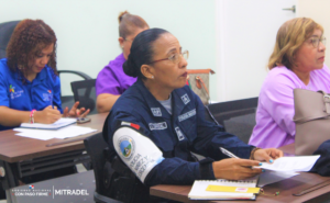 A group of women, including a uniformed officer, are attentively listening in a classroom setting, taking notes.